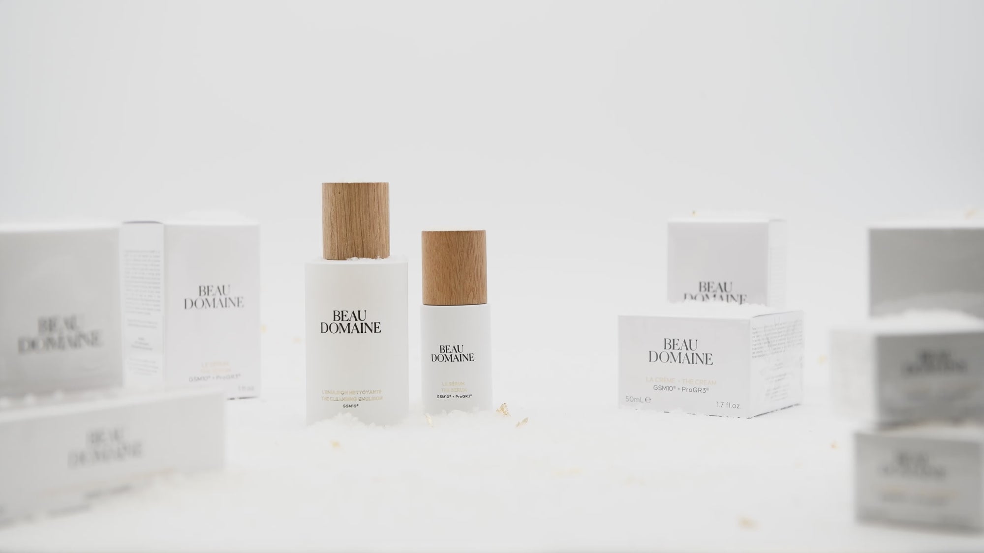 Static video showing an animation of La Routine Complète in the world of holiday celebrations, with products lined up against a clean white background and fake snow, with a hand applying cream in the foreground